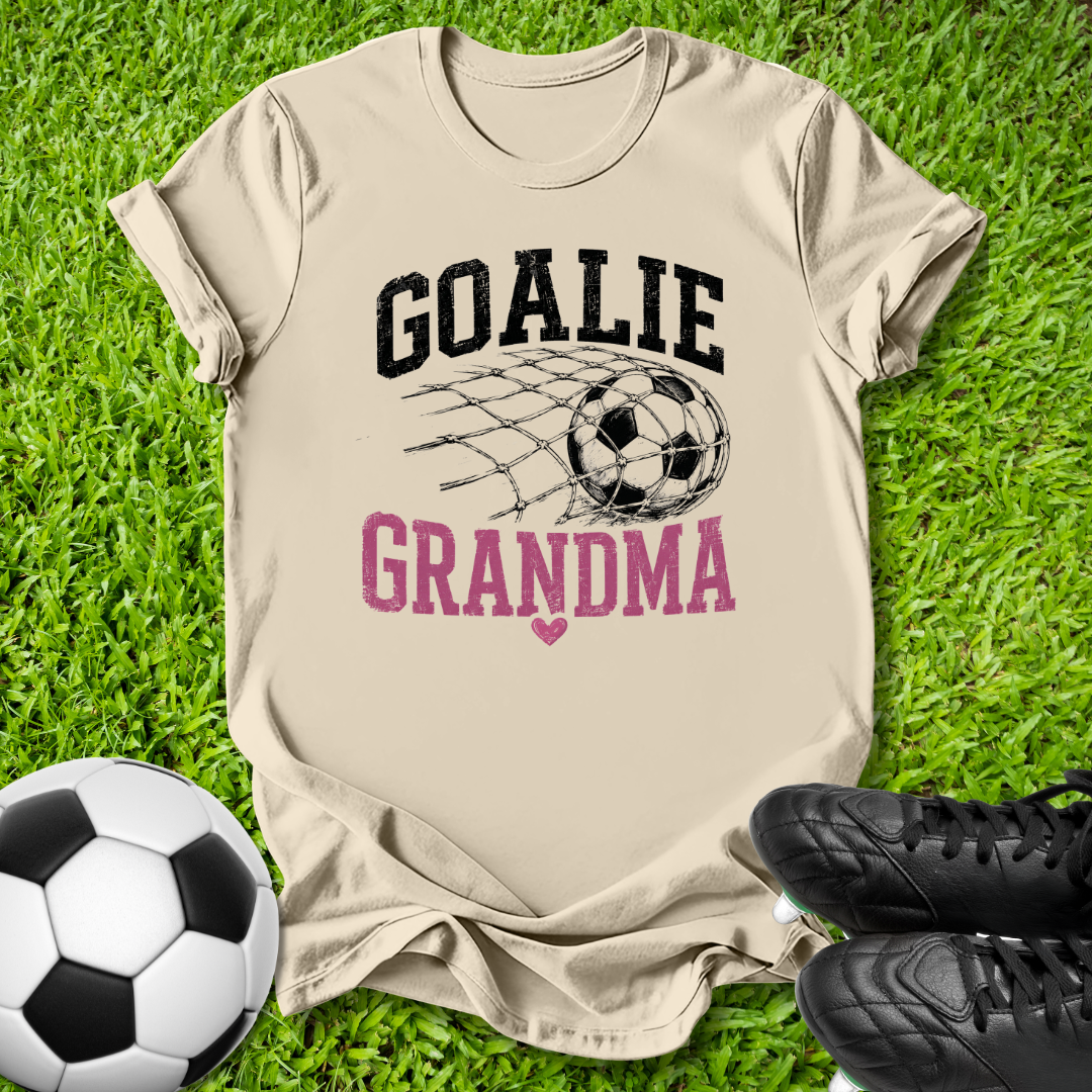 A beige graphic t-shirt laid flat on green grass, featuring the words 'GOALIE GRANDMA' with a soccer ball and net design. A soccer ball and black cleats are placed around it as props.