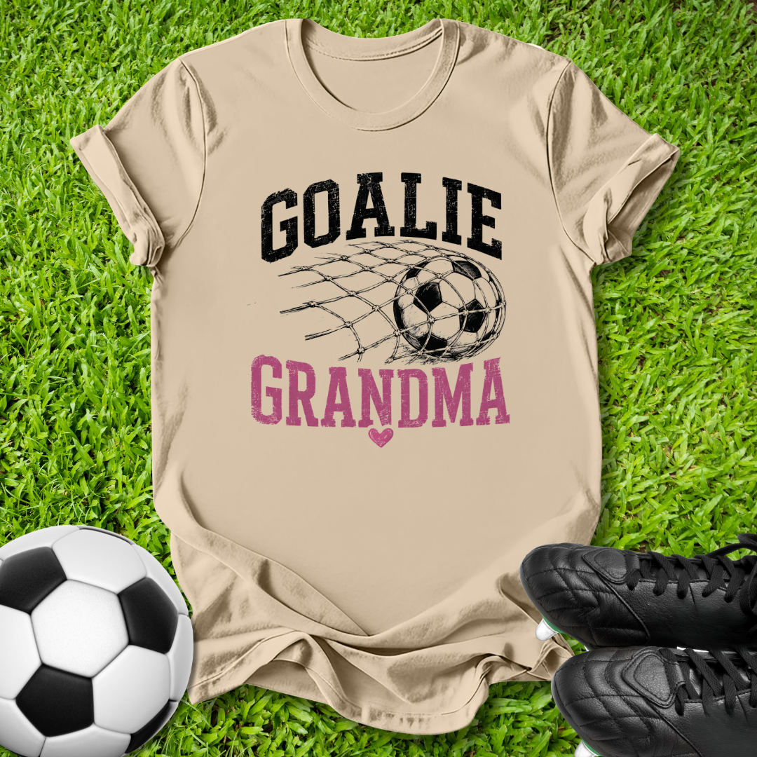 A beige graphic t-shirt laid flat on green grass, featuring the words 'GOALIE GRANDMA' with a soccer ball and net design. A soccer ball and black cleats are placed around it as props.