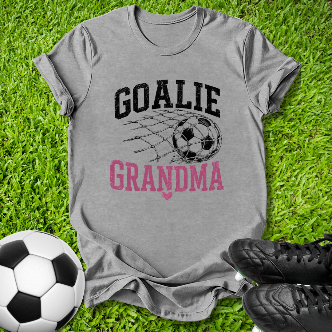 A heather gray graphic t-shirt laid flat on green grass, featuring the words 'GOALIE GRANDMA' in black and pink with a soccer ball and net design. A soccer ball and black cleats are placed around it as props.