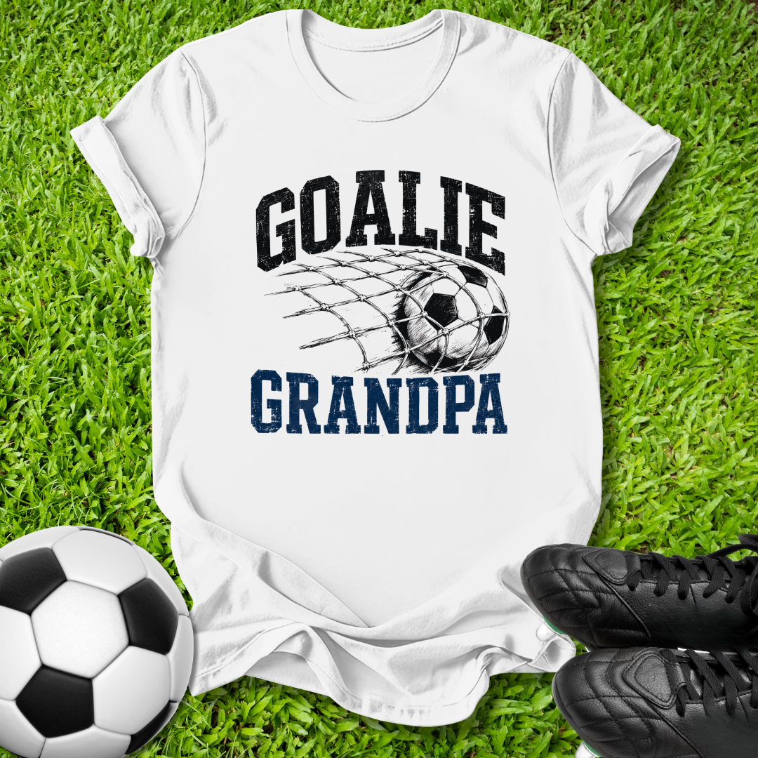white graphic t-shirt laid flat on green grass, featuring a soccer net design with the words 'GOALIE GRANDPA' in black and blue. A soccer ball and black cleats are styled around the shirt.