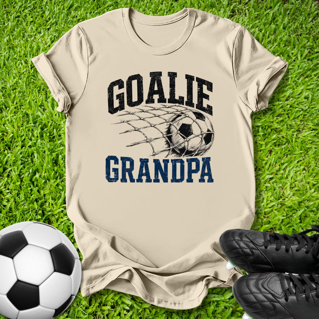 A beige graphic t-shirt laid flat on green grass, featuring the text 'GOALIE GRANDPA' with a soccer net and ball design. A soccer ball and black cleats are placed around it as props.
