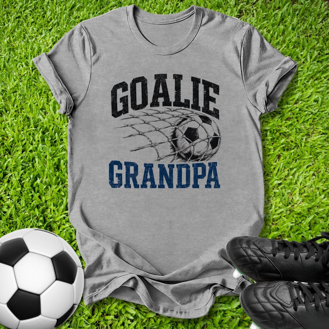 A heather gray graphic t-shirt laid flat on green grass, featuring a soccer net design with the words 'GOALIE GRANDPA' in black and blue. A soccer ball and black cleats are styled around the shirt.