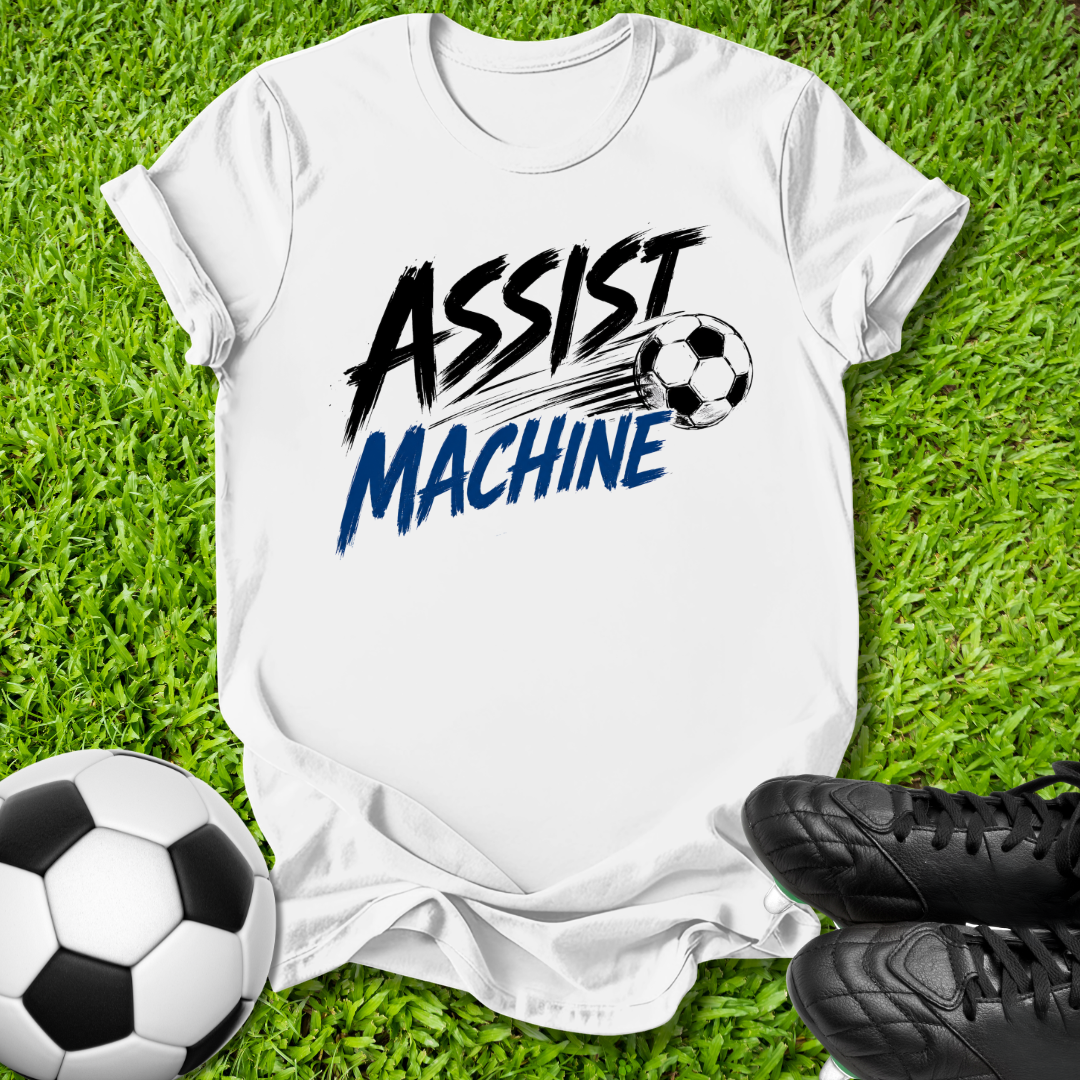 A white short-sleeve graphic T-shirt laid flat on green grass, featuring the words 'ASSIST MACHINE' in black and blue with a soccer ball illustration. A black-and-white soccer ball and black cleats are placed around it as props.