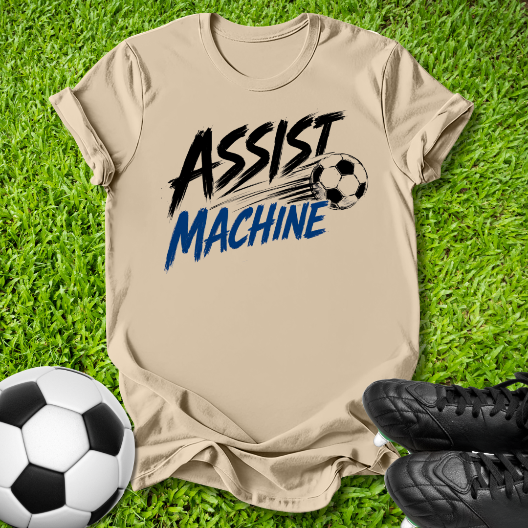A beige graphic t-shirt laid flat on green grass, featuring the words 'ASSIST MACHINE' in black and blue with a soccer ball illustration. A soccer ball and black cleats are placed around it as props.