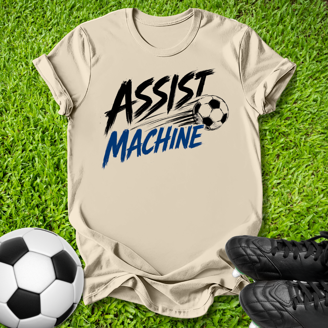 A beige graphic t-shirt laid flat on green grass, featuring the words 'ASSIST MACHINE' in black and blue brush-style lettering with a soccer ball illustration. A soccer ball and black cleats are placed around it as props.