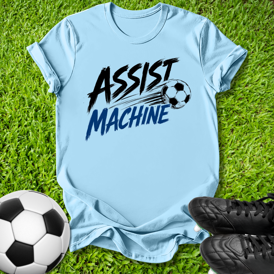 A light blue graphic t-shirt laid flat on green grass, featuring the text 'ASSIST MACHINE' in black and blue with a soccer ball illustration. A soccer ball and black cleats are placed around it as props.