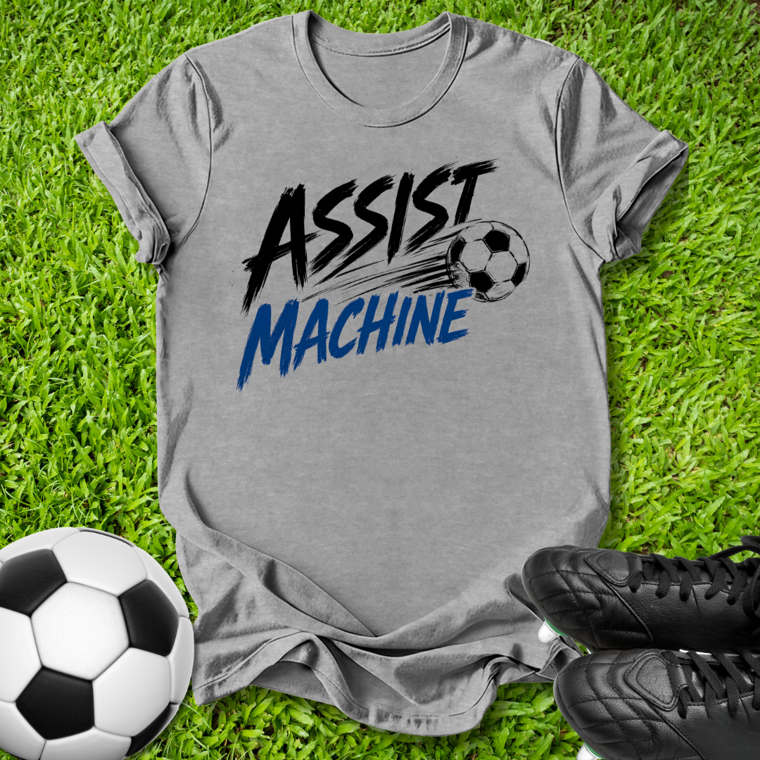 A heather gray graphic t-shirt laid flat on green grass, featuring the words 'ASSIST MACHINE' in black and blue with a soccer ball illustration. A soccer ball and black cleats are placed around it as props.