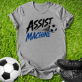 A heather gray graphic t-shirt laid flat on green grass, featuring the words 'ASSIST MACHINE' in black and blue with a soccer ball illustration. A soccer ball and black cleats are placed around it as props.