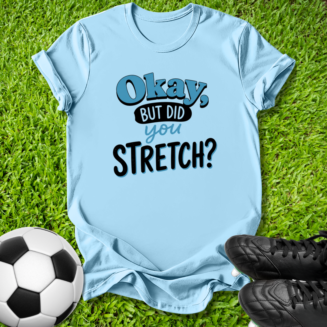 A light blue graphic t-shirt laid flat on green grass, featuring the text 'Okay, BUT DID you STRETCH?' in blue and black. A soccer ball and black cleats are placed at the bottom corners as props.