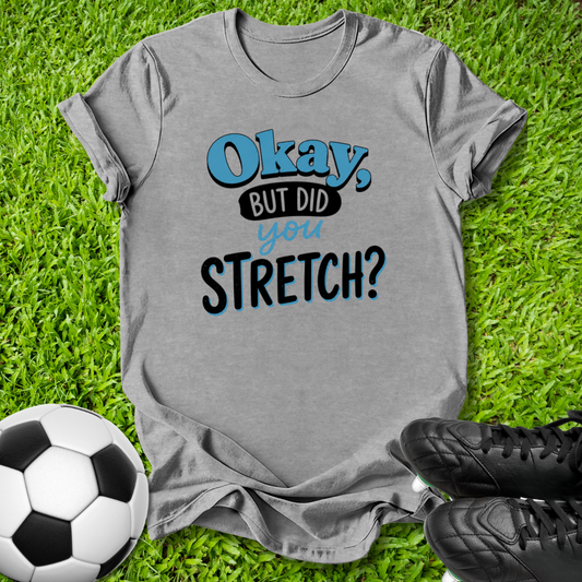 A heather gray graphic t-shirt laid flat on green grass, featuring the text 'Okay, BUT DID you STRETCH?' in blue and black. A soccer ball is visible on the bottom left and black cleats are on the bottom right.