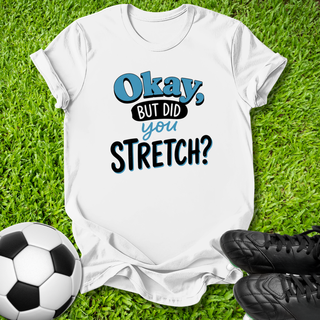 A white graphic t-shirt laid flat on green grass, featuring the text 'Okay, BUT DID you STRETCH?' in blue and black lettering. A soccer ball is visible in the bottom-left corner and a pair of black cleats is visible in the bottom-right corner.
