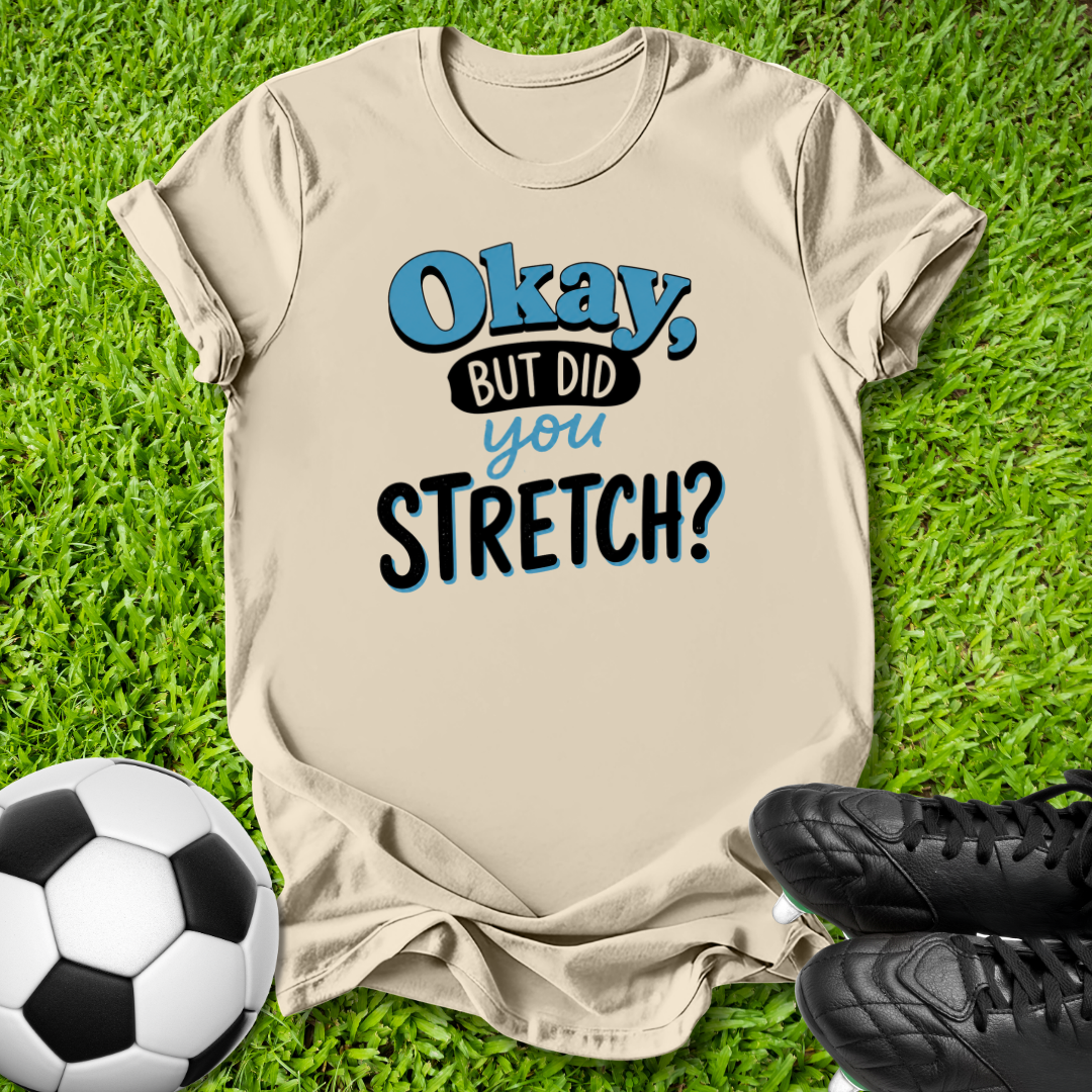 A beige graphic t-shirt laid flat on green grass with a soccer ball and black cleats as props. The shirt features the text 'Okay, BUT DID you STRETCH?' in blue and black lettering.