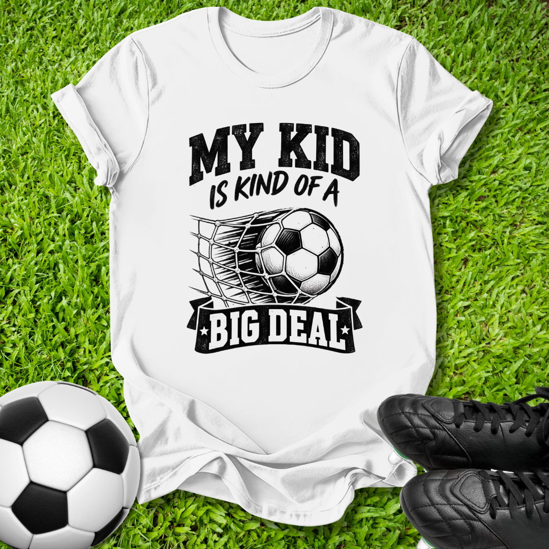 A white short-sleeve graphic T-shirt laid flat on green grass, with a soccer ball and black cleats nearby. The shirt has a black printed slogan reading 'MY KID IS KIND OF A BIG DEAL' around a soccer ball and net graphic.