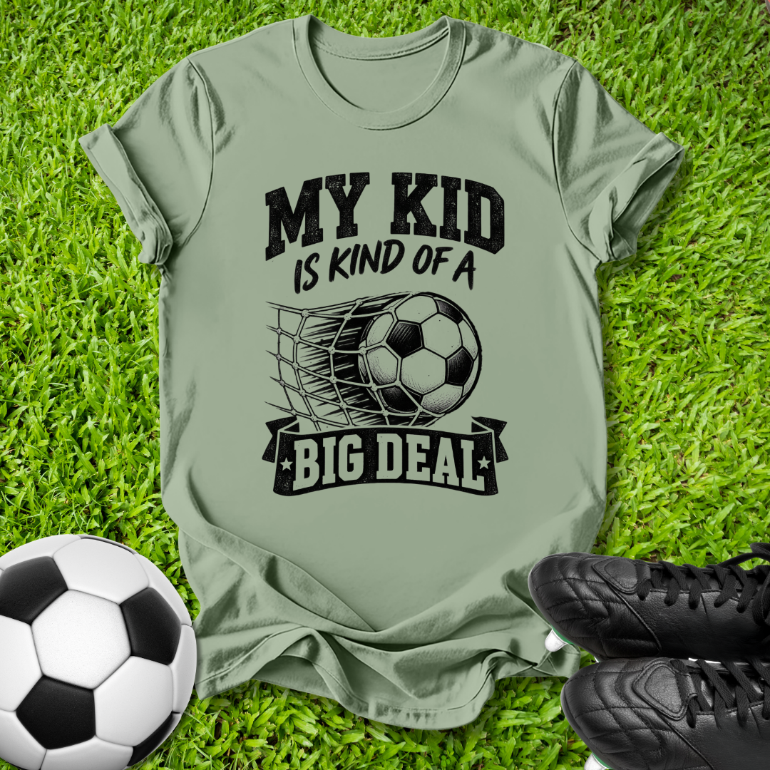 An olive green graphic t-shirt laid flat on green grass, featuring a black print that reads 'MY KID IS KIND OF A BIG DEAL' with a soccer ball and net graphic. A soccer ball and black cleats are placed around it as props.