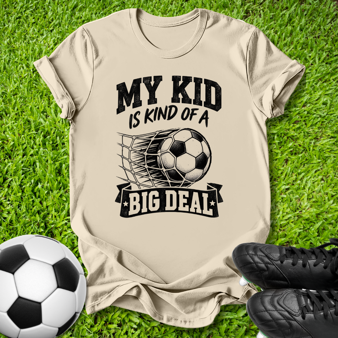 A beige graphic t-shirt laid flat on green grass, featuring a black print that reads 'MY KID IS KIND OF A BIG DEAL' with a soccer ball and net graphic. A soccer ball and black cleats are placed around it as props.