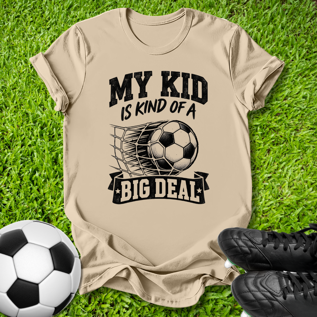 A beige graphic t-shirt laid flat on green grass, featuring a black print that reads 'MY KID IS KIND OF A BIG DEAL' with a soccer ball and net graphic. A soccer ball and black cleats are placed around it as props.