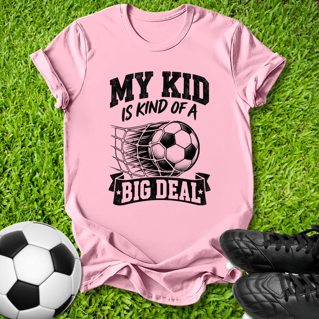 A pink short-sleeve t-shirt with a black graphic reading 'MY KID IS KIND OF A BIG DEAL' around a soccer ball and net illustration, laid flat on green grass with a soccer ball and black cleats nearby.