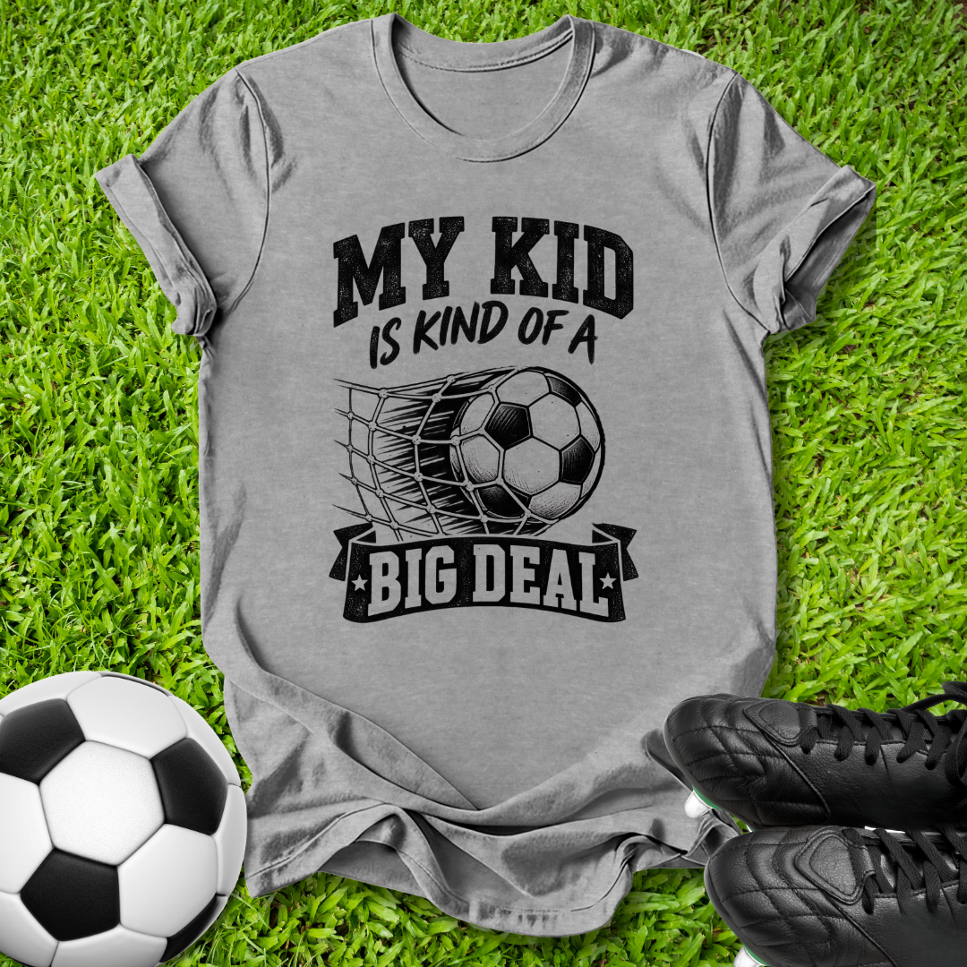 A heather gray graphic t-shirt laid flat on green grass, featuring a black print that reads 'MY KID IS KIND OF A BIG DEAL' with a soccer ball and net graphic. A soccer ball and black cleats are styled around the shirt.