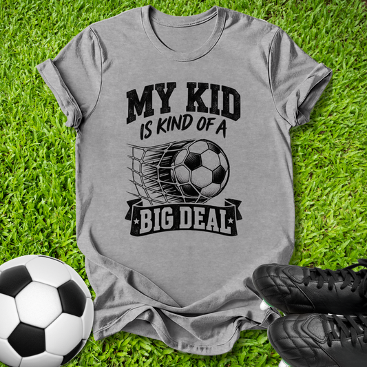 A heather gray graphic t-shirt laid flat on green grass, featuring a black print that reads 'MY KID IS KIND OF A BIG DEAL' with a soccer ball and net graphic. A soccer ball and black cleats are styled around the shirt.