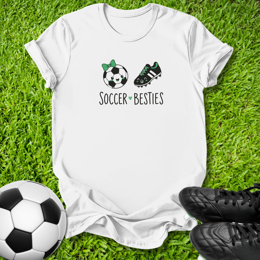 A white short-sleeve T-shirt laid flat on green grass, featuring a soccer-themed graphic with a soccer ball, a cleat, and the words 'SOCCER BESTIES'. A black-and-white soccer ball appears at the lower left, and black cleats are at the lower right.