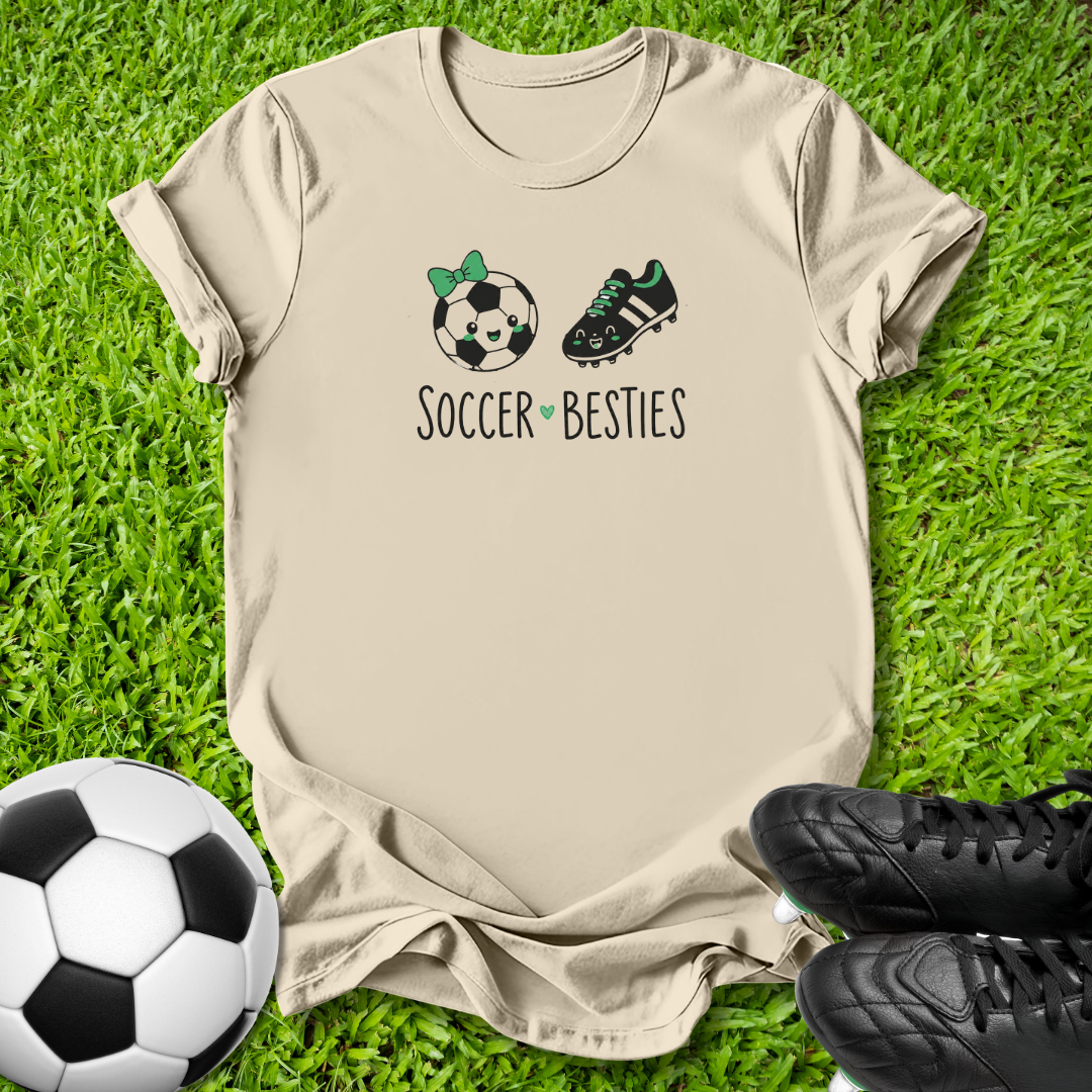 A beige graphic t-shirt laid flat on green grass, featuring a soccer ball and cleat illustration with the text 'SOCCER BESTIES'. A soccer ball and black cleats are placed around it as props.
