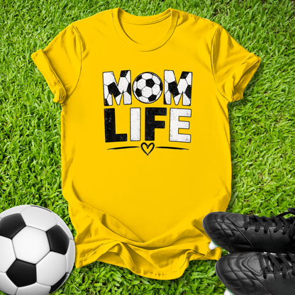 A bright yellow graphic t-shirt laid flat on green grass, featuring the words 'MOM LIFE' in black and white soccer-ball-patterned lettering. A soccer ball and black cleats are styled around the shirt.