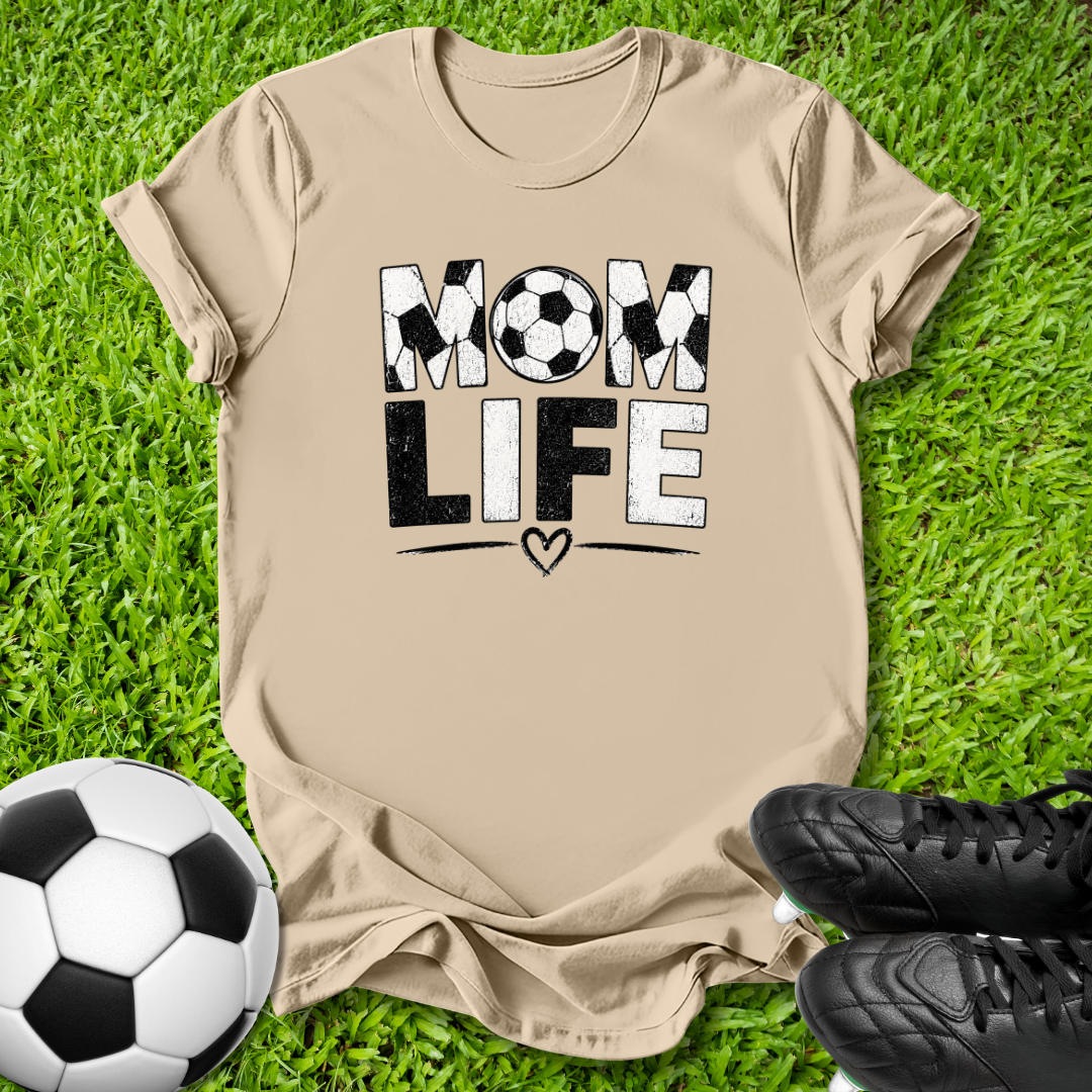 A beige graphic t-shirt laid flat on green grass, featuring the words 'MOM LIFE' in black and white with soccer ball and heart details. A soccer ball and black cleats are placed around it as props.
