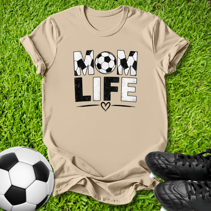 A beige graphic t-shirt laid flat on green grass, featuring the words 'MOM LIFE' in black and white with soccer ball and heart details. A soccer ball and black cleats are placed around it as props.