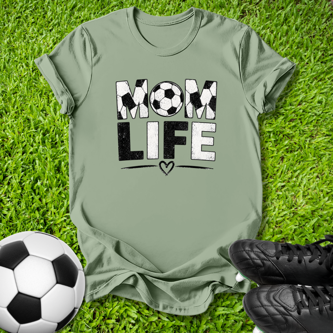 An olive green t-shirt laid flat on green grass, featuring a 'MOM LIFE' graphic with soccer ball patterns. A soccer ball and black cleats are styled around it.