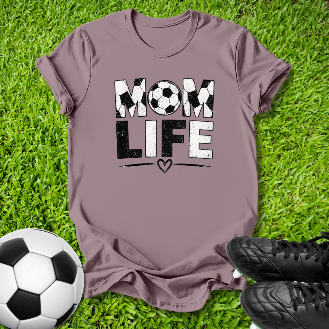 A mauve short-sleeve t-shirt laid flat on green grass, featuring a distressed graphic that reads 'MOM LIFE' with soccer ball patterns inside the letters. A soccer ball and black cleats are placed around it as props.