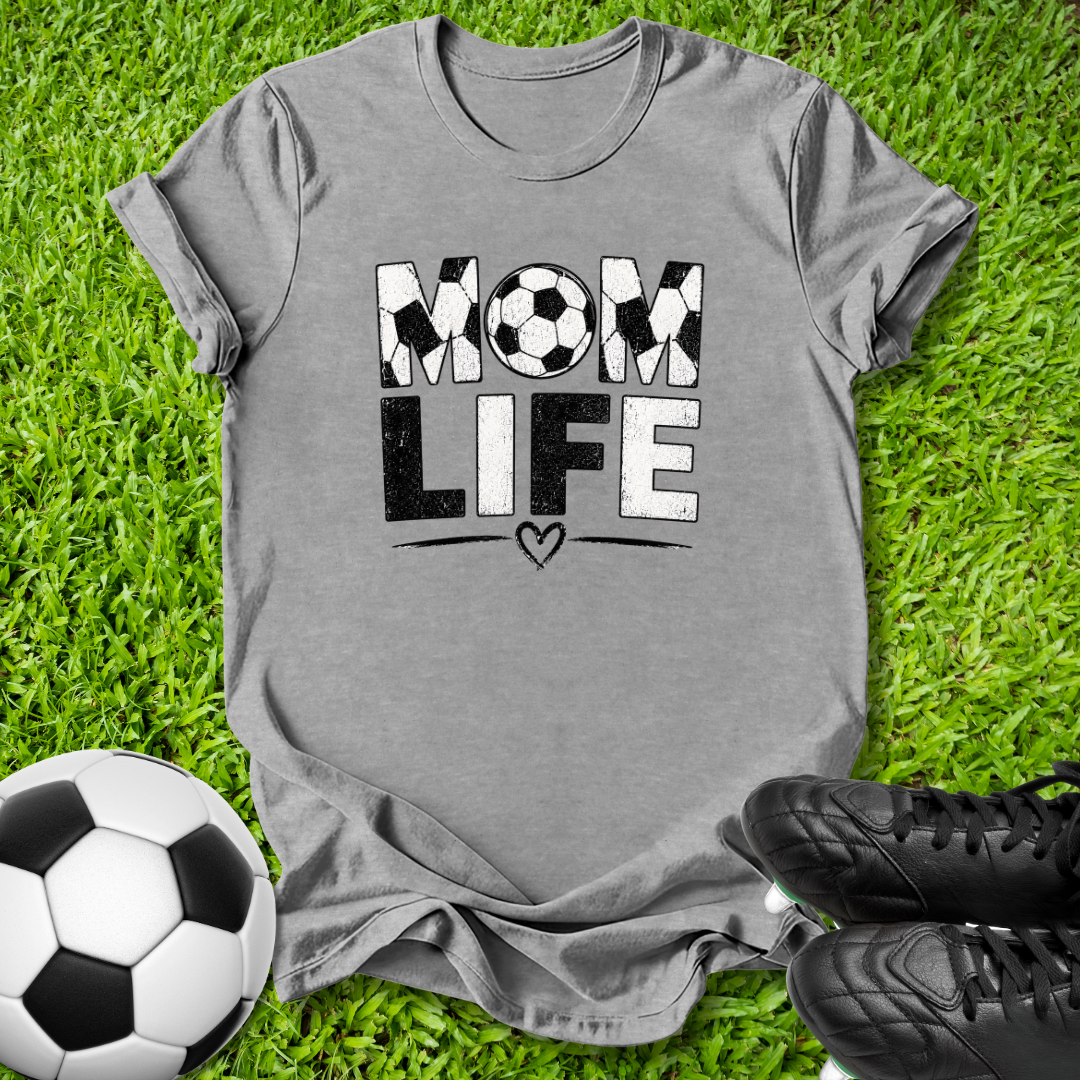 A heather gray short-sleeve t-shirt with a 'MOM LIFE' graphic printed on the front, laid flat on green grass with a soccer ball and black cleats as props.