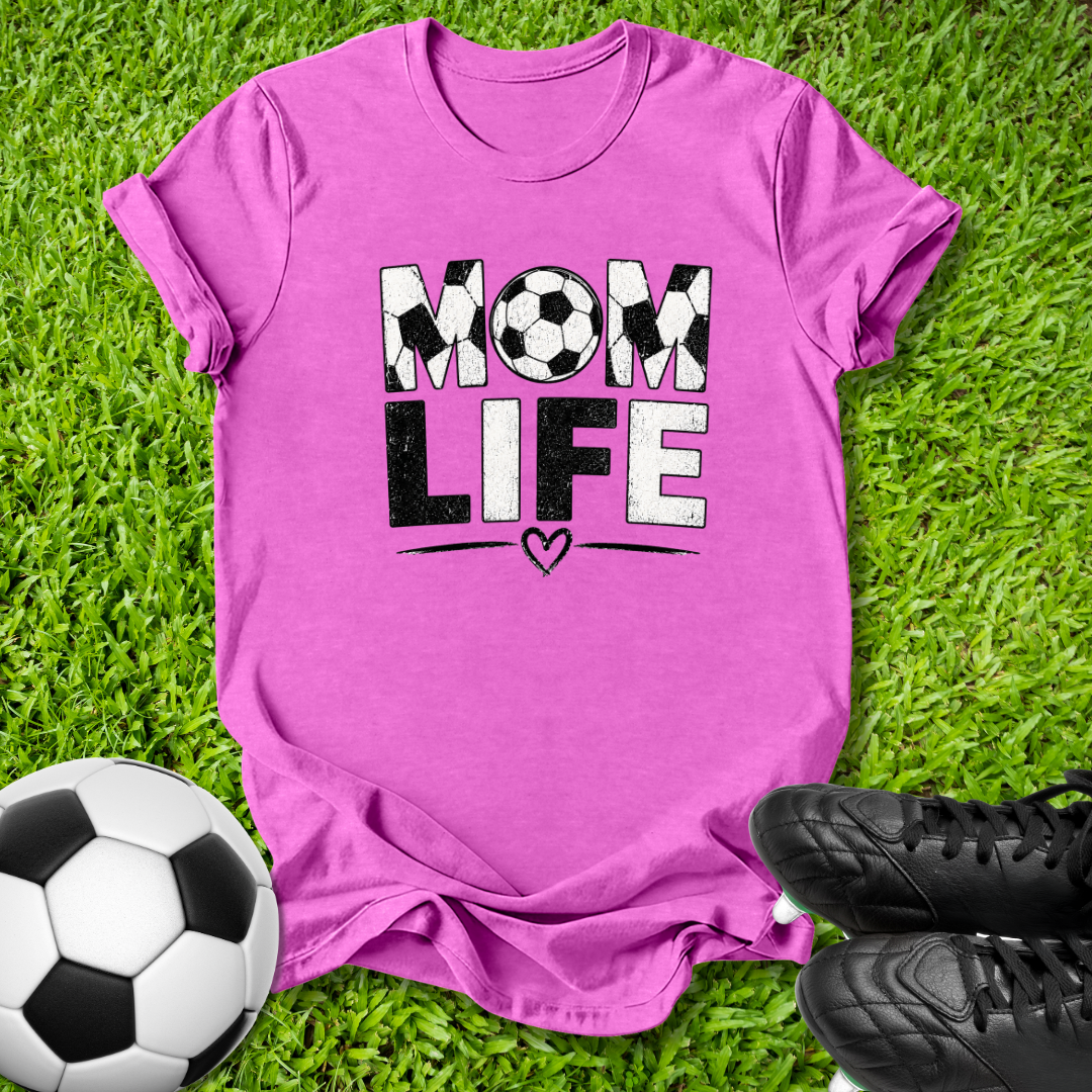 A bright pink short-sleeve t-shirt laid flat on green grass, featuring the words 'MOM LIFE' in black and white with soccer ball patterns. A soccer ball is on the bottom left, and black cleats are on the bottom right.