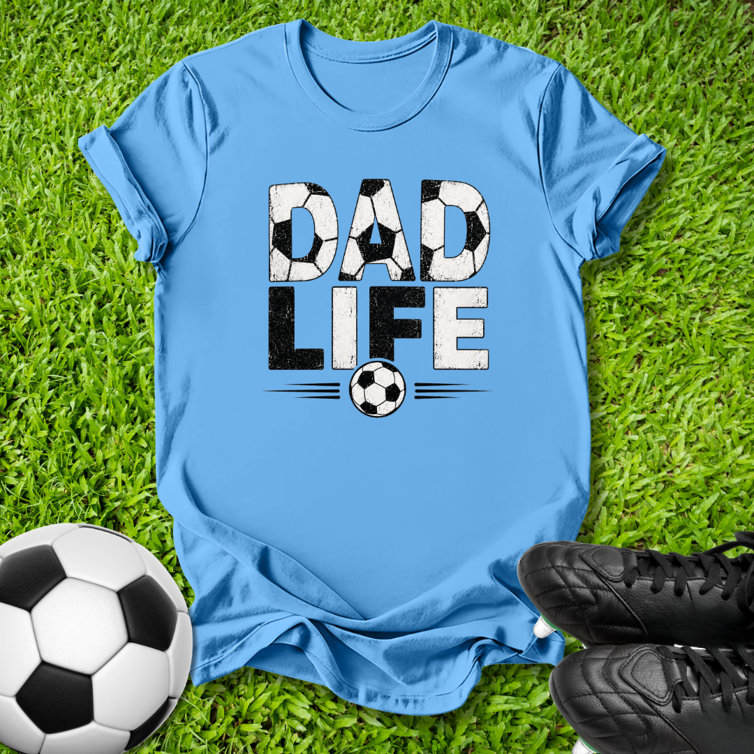 A light blue crewneck t-shirt laid flat on green grass, featuring a graphic that reads 'DAD LIFE' with soccer ball patterns inside the letters and a soccer ball below. A real soccer ball and black cleats are placed around it as props.