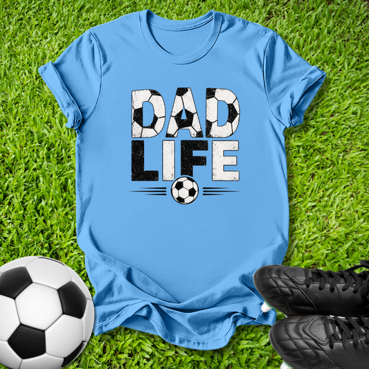 A light blue crewneck t-shirt laid flat on green grass, featuring a graphic that reads 'DAD LIFE' with soccer ball patterns inside the letters and a soccer ball below. A real soccer ball and black cleats are placed around it as props.