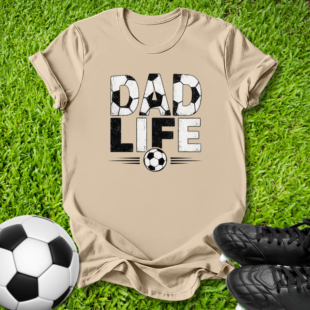 A beige graphic t-shirt laid flat on green grass, featuring the words 'DAD LIFE' in a soccer-ball-themed design. A soccer ball and black cleats are styled around the shirt.
