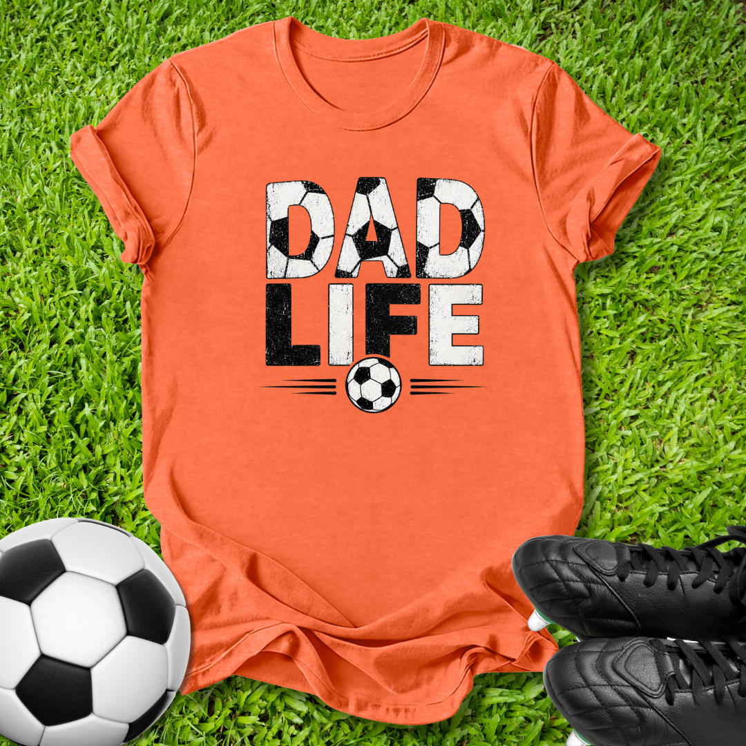 An orange graphic t-shirt laid flat on green grass, featuring the words 'DAD LIFE' in a soccer-themed design. A soccer ball is visible in the bottom-left corner and a pair of black soccer cleats is visible in the bottom-right corner.