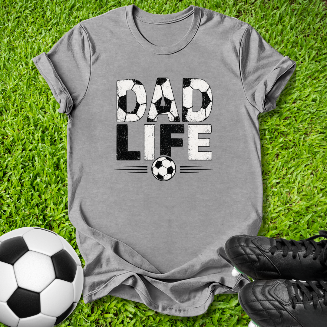 A heather gray graphic t-shirt laid flat on green grass, featuring a distressed black and white soccer-themed 'DAD LIFE' print. A soccer ball and black cleats are styled around the shirt.