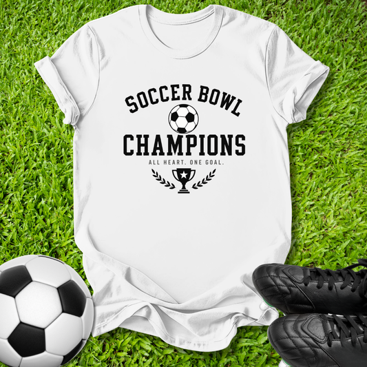 A white short-sleeve T-shirt laid flat on green grass, printed with the words 'SOCCER BOWL CHAMPIONS' and a trophy graphic in black. A soccer ball is at the lower left, and black cleats are at the lower right.