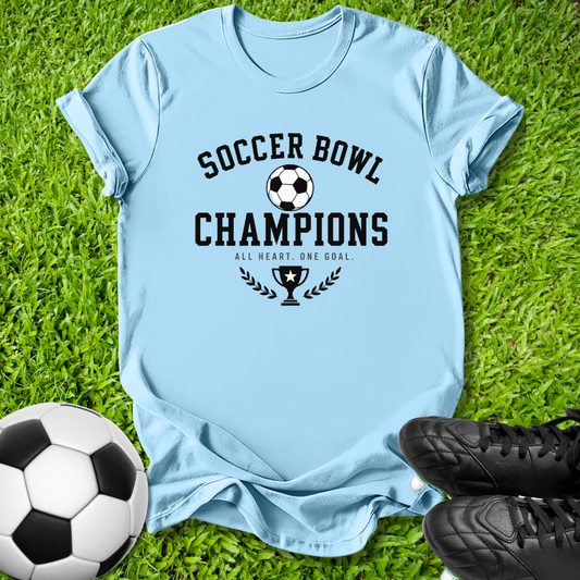 A light blue graphic t-shirt laid flat on green grass, featuring a black 'SOCCER BOWL CHAMPIONS' design with a soccer ball and trophy graphic. A soccer ball and black cleats are placed around it as props.