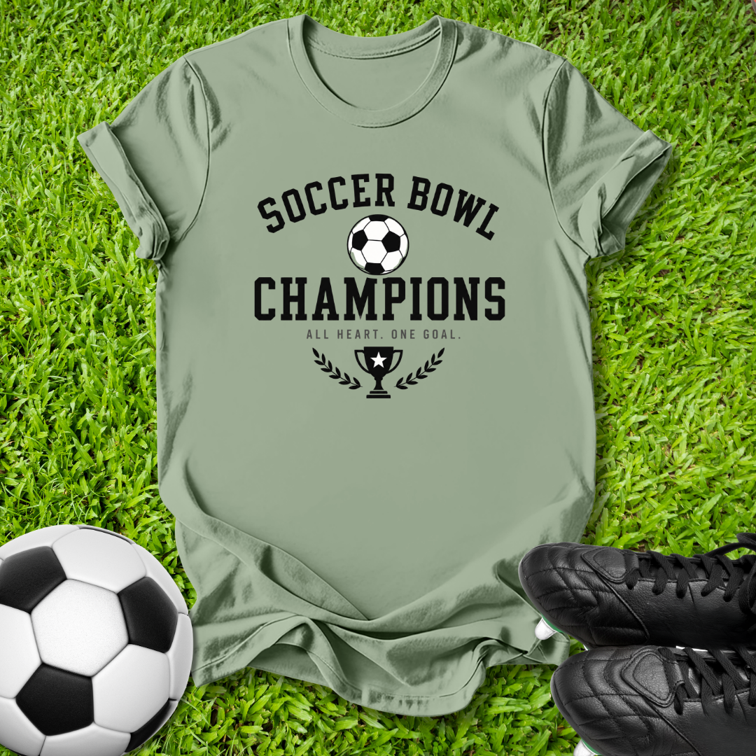 An olive green t-shirt with a black 'Soccer Bowl Champions' graphic, laid flat on green grass with a soccer ball and black cleats as props.