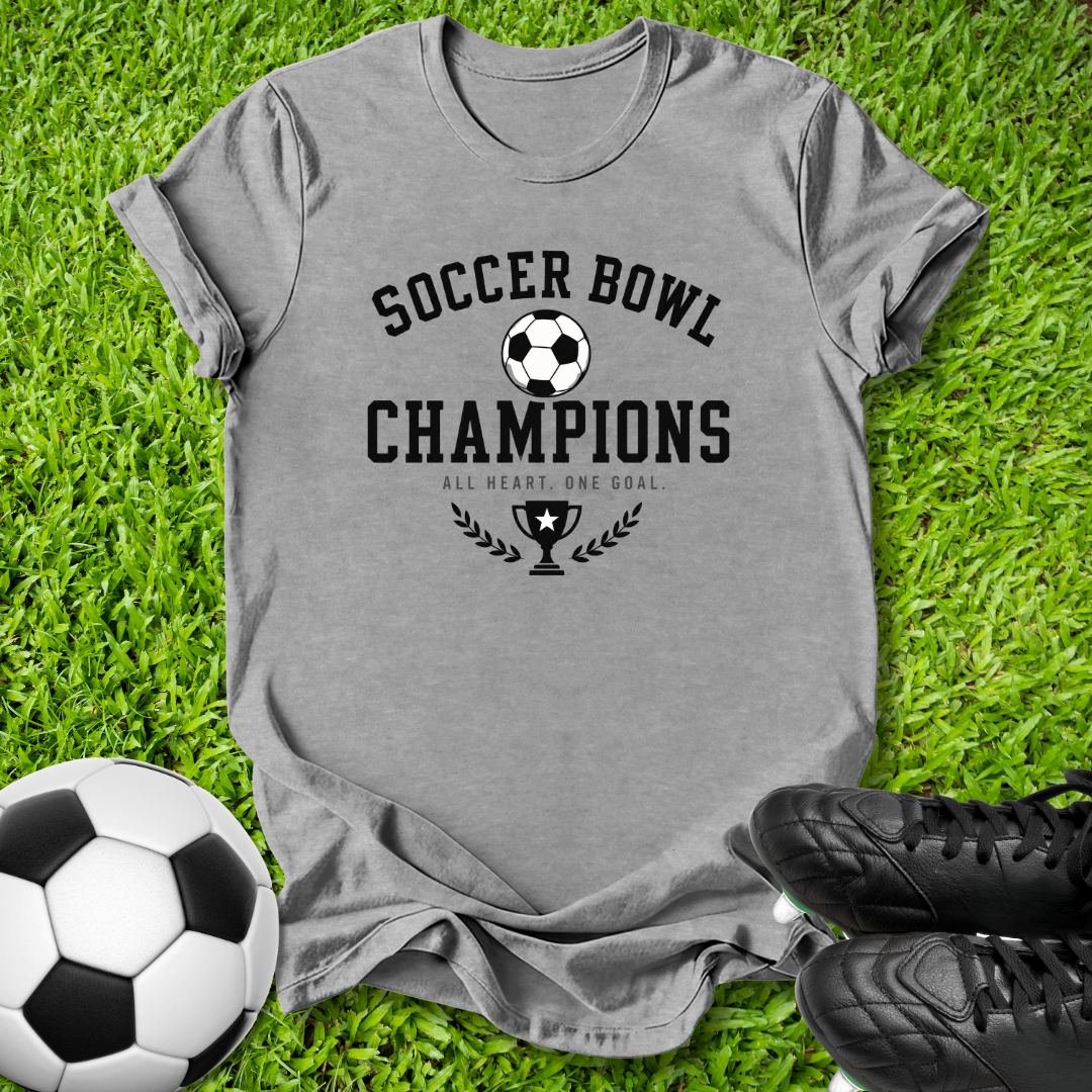 A heather gray short-sleeve t-shirt with a black 'Soccer Bowl Champions' graphic, laid flat on green grass with a soccer ball and black cleats nearby.