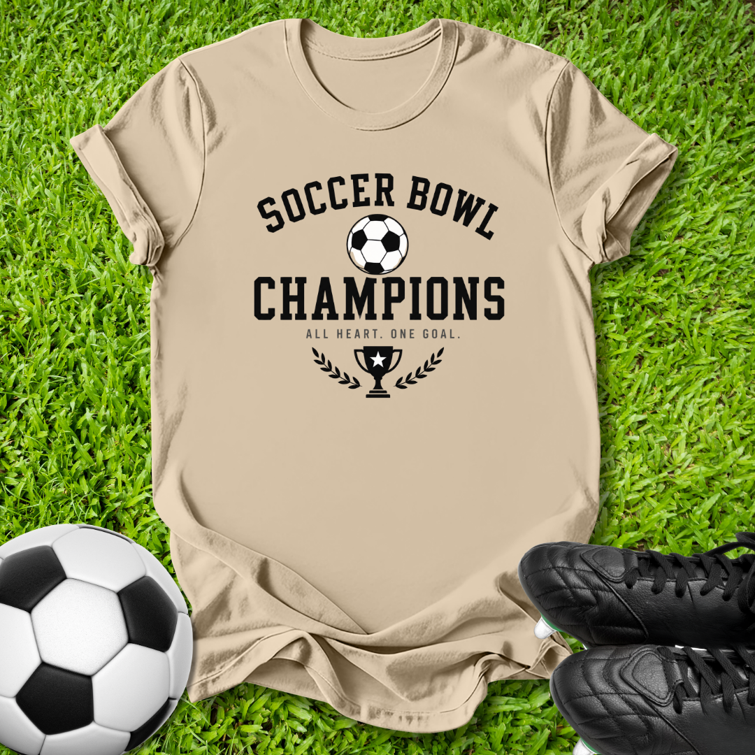 A beige graphic t-shirt laid flat on green grass, featuring a black soccer-themed print that reads 'SOCCER BOWL CHAMPIONS' with a trophy and laurel design. A soccer ball is on the left and black cleats are on the right.