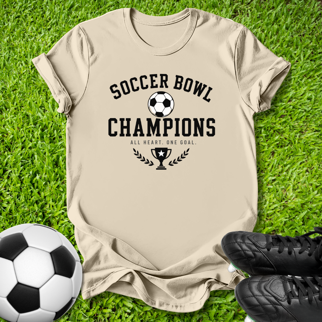 A beige graphic t-shirt laid flat on green grass, featuring a black soccer-themed print that reads 'SOCCER BOWL CHAMPIONS' with a trophy and laurel design. A soccer ball is at the bottom left, and black cleats are at the bottom right.