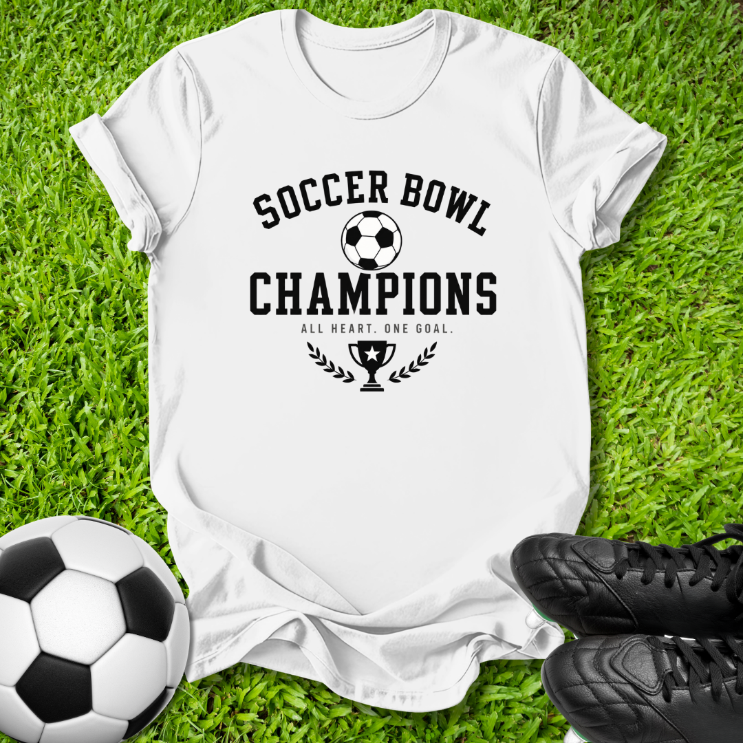 A white short-sleeve T-shirt laid flat on green grass, printed with the words 'SOCCER BOWL CHAMPIONS' and a trophy graphic in black. A soccer ball is at the lower left, and black cleats are at the lower right.