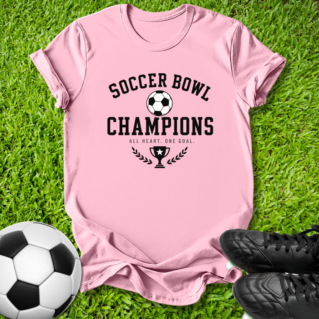 A light pink short-sleeve t-shirt with a black graphic reading 'SOCCER BOWL CHAMPIONS' and 'ALL HEART, ONE GOAL' with a trophy and soccer ball motif, laid flat on green grass with a soccer ball and black cleats nearby.