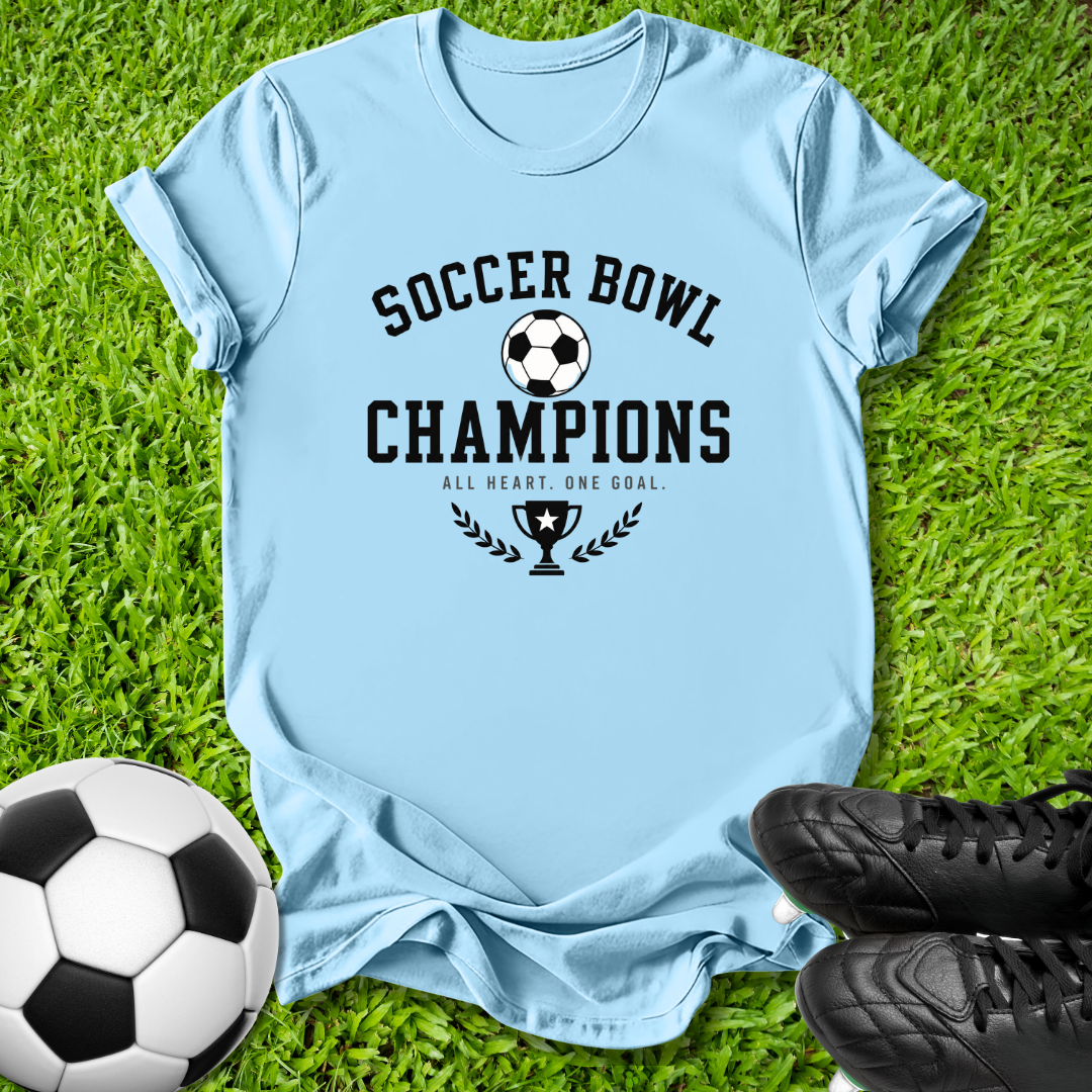 A light blue graphic t-shirt laid flat on green grass, featuring a black 'SOCCER BOWL CHAMPIONS' design with a soccer ball and trophy graphic. A soccer ball and black cleats are placed around it as props.