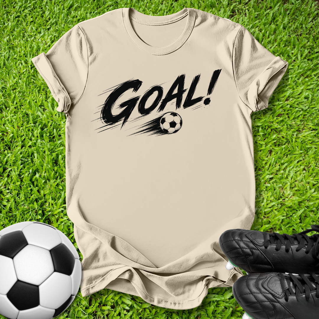 A beige short-sleeve t-shirt with a black 'GOAL!' graphic and soccer ball print, laid flat on green grass with a soccer ball and black cleats as props.
