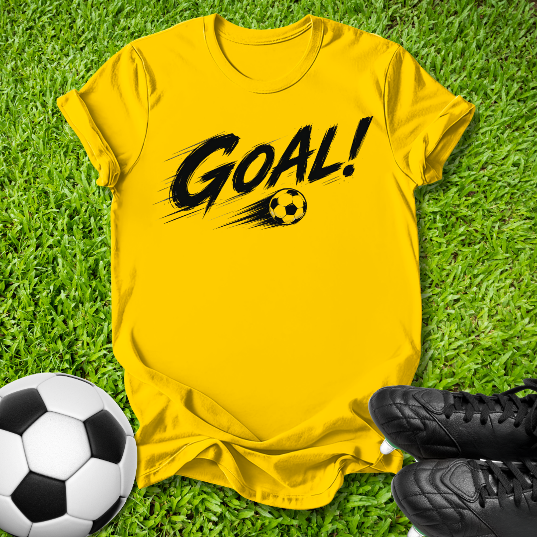 A bright yellow short-sleeve t-shirt with a black 'GOAL!' graphic and soccer ball print, laid flat on green grass with a soccer ball and black cleats nearby.