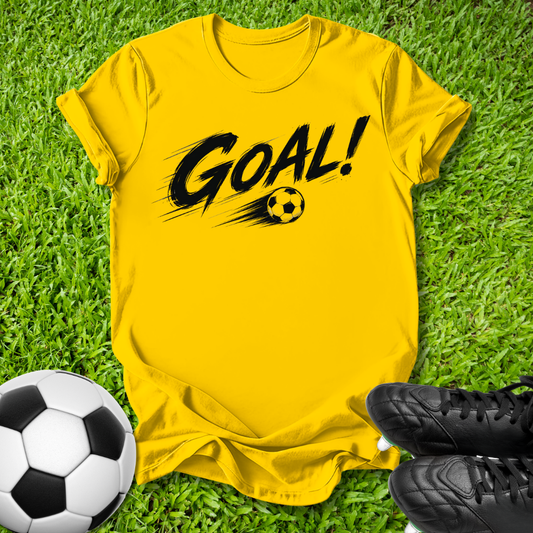 A bright yellow short-sleeve t-shirt with a black 'GOAL!' graphic and soccer ball print, laid flat on green grass with a soccer ball and black cleats nearby.
