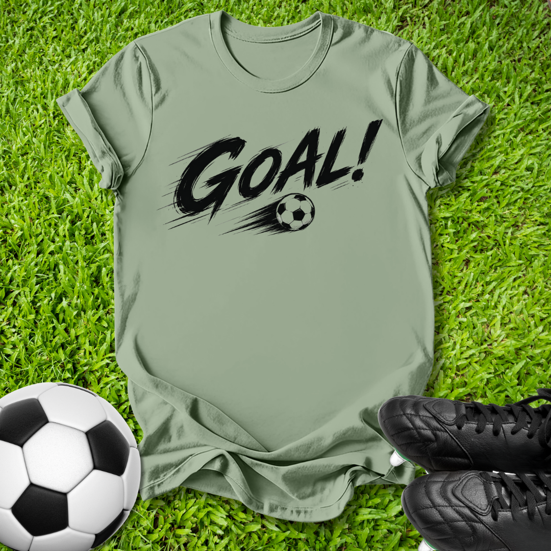 An olive green short-sleeve t-shirt with a black 'GOAL!' graphic and soccer ball print, laid flat on green grass with a soccer ball and black cleats as props.